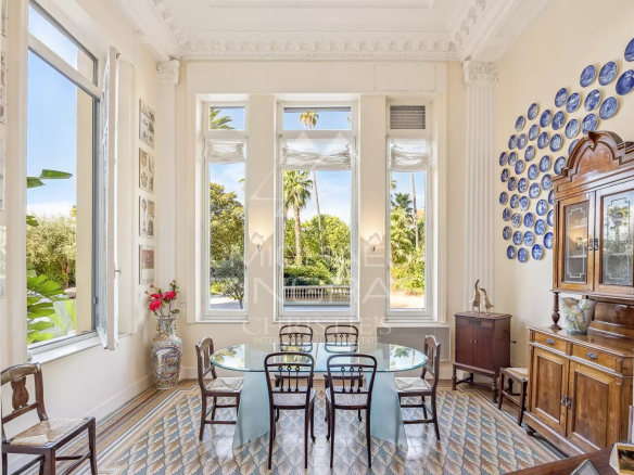 Bright dining room with a glass table, six wooden chairs, and tall windows overlooking greenery; blue plate wall decor on the right.