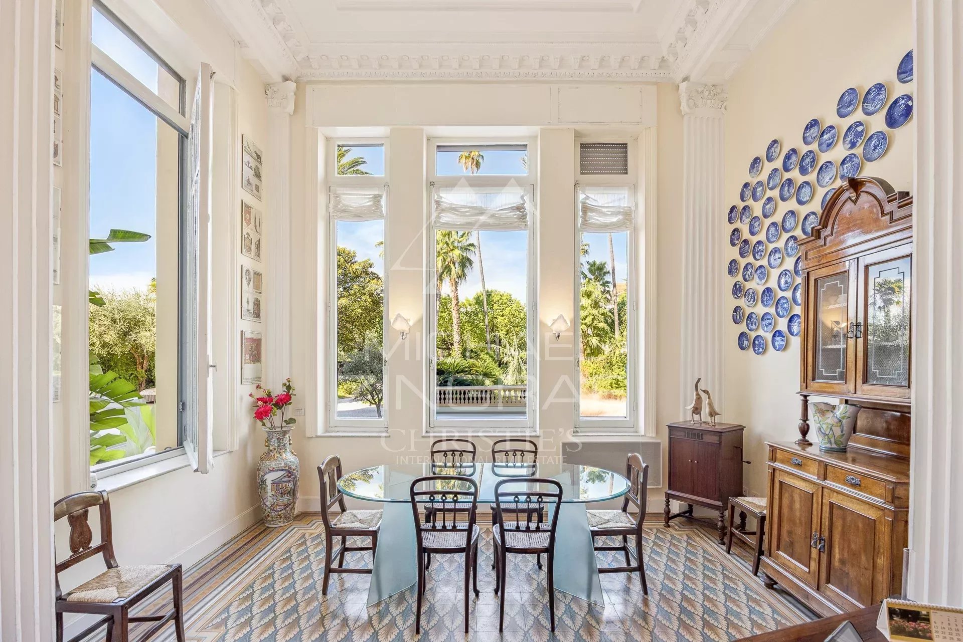 Bright dining room with a glass table, six wooden chairs, and tall windows overlooking greenery; blue plate wall decor on the right.