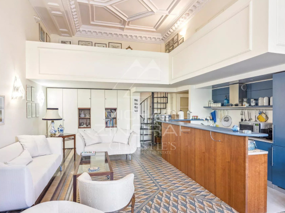 Open-plan living area with white sofas, glass coffee table, built-in cabinets, and a blue-and-wood kitchen bar under a detailed white ceiling with decorative panels; patterned tile floor.