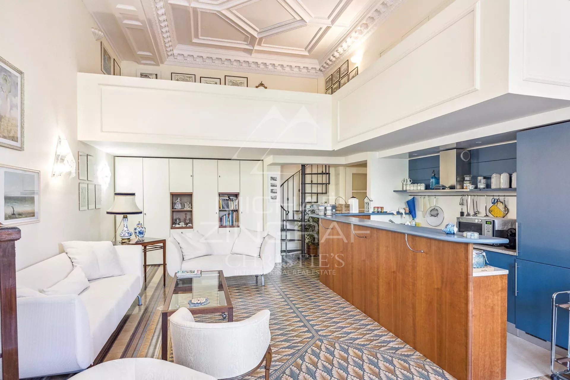 Open-plan living area with white sofas, glass coffee table, built-in cabinets, and a blue-and-wood kitchen bar under a detailed white ceiling with decorative panels; patterned tile floor.