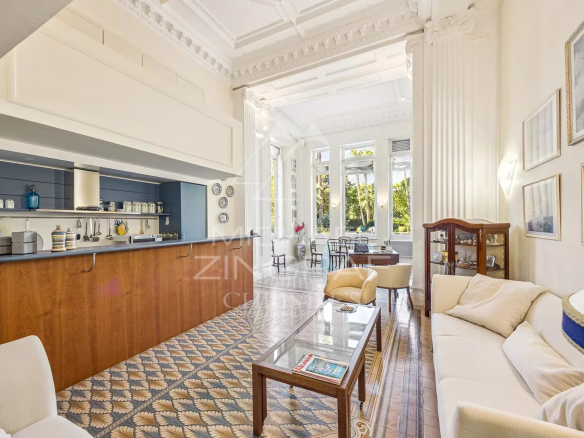 Open-plan living room with a wooden bar counter on the left, white ornate ceiling, and large windows letting in greenery; seating area with a glass coffee table and cream sofa.