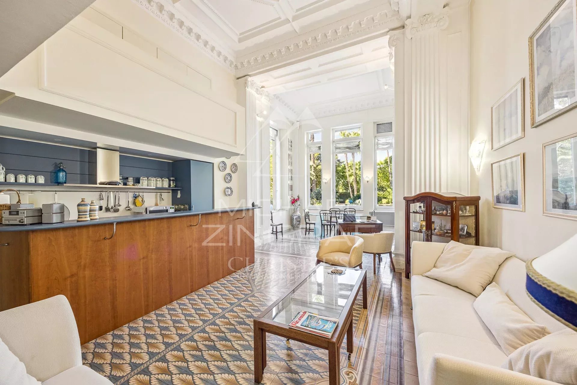 Open-plan living room with a wooden bar counter on the left, white ornate ceiling, and large windows letting in greenery; seating area with a glass coffee table and cream sofa.