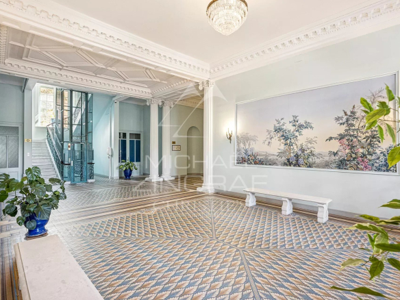 Elegant hotel lobby with an ornate white ceiling, chandelier, and pale blue walls, featuring a patterned tile floor and a staircase nearby.