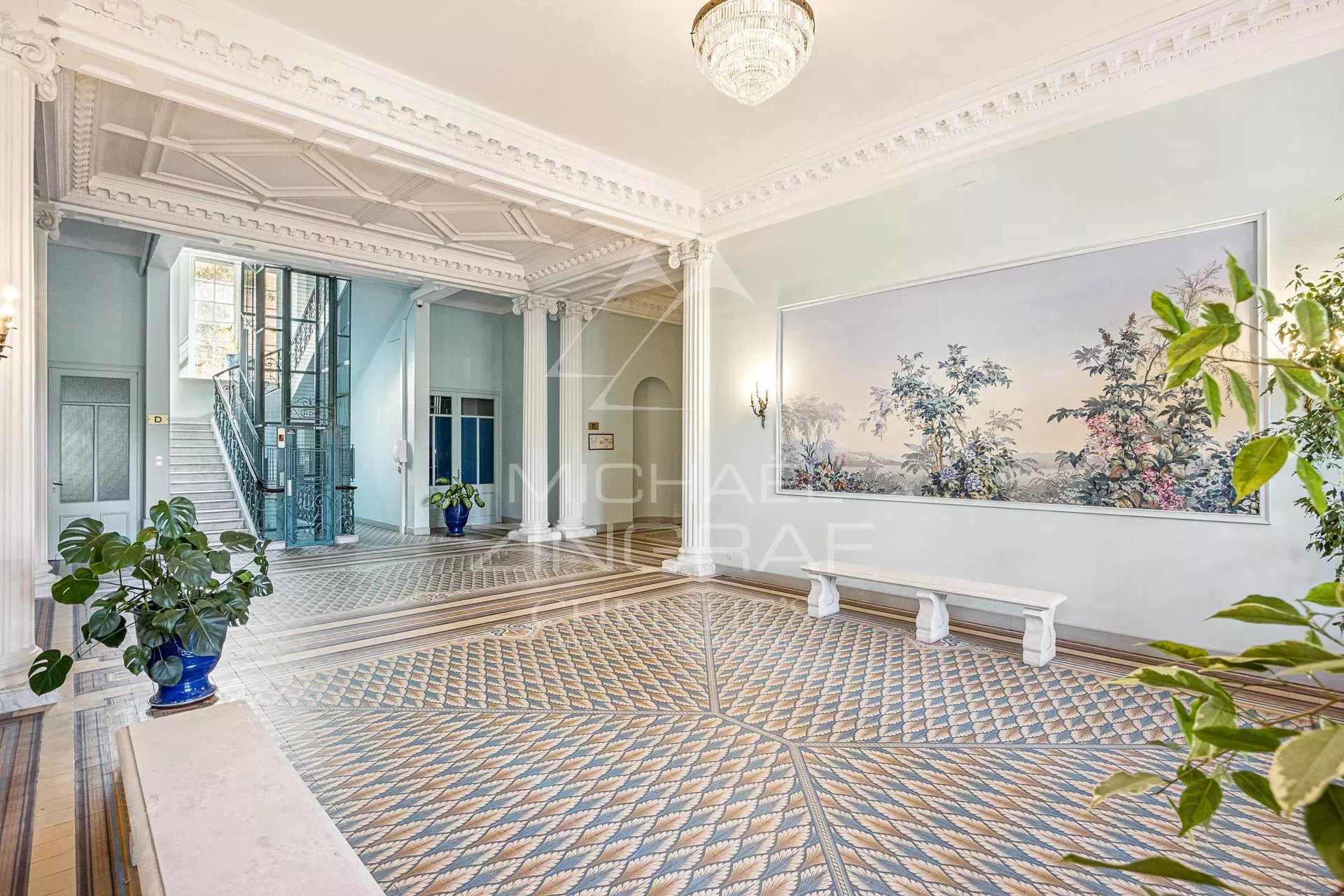Elegant hotel lobby with an ornate white ceiling, chandelier, and pale blue walls, featuring a patterned tile floor and a staircase nearby.