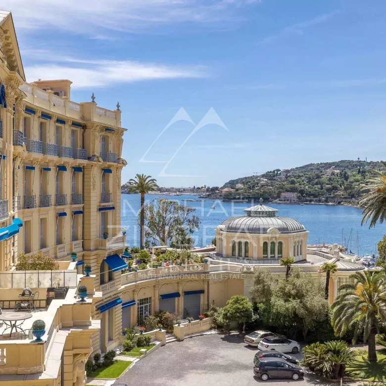 Four-story Mediterranean hotel with cream-yellow walls, blue awnings, and ornate balconies overlooking a courtyard and the blue sea beyond.