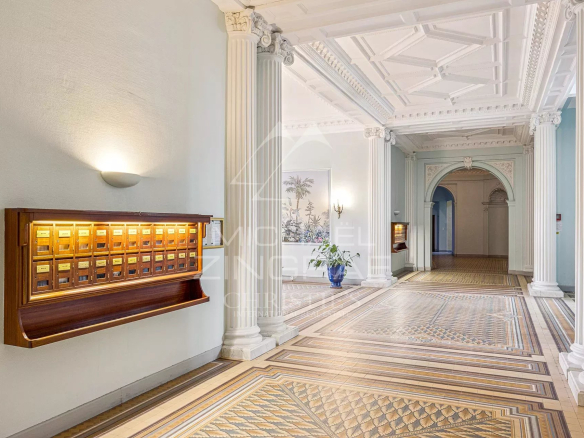 Elegant hotel lobby interior with tall decorative columns, an ornate coffered ceiling, and patterned tile floor; a wooden mailbox unit is mounted on the left wall.