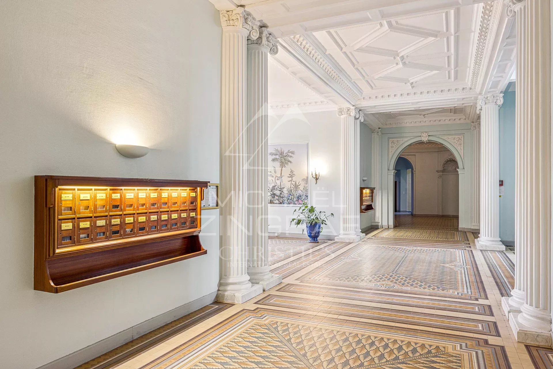 Elegant hotel lobby interior with tall decorative columns, an ornate coffered ceiling, and patterned tile floor; a wooden mailbox unit is mounted on the left wall.