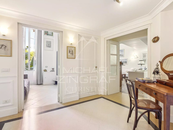 Bright hallway with a wooden dressing table and round mirror on the right, two open doorways leading to bedrooms and living area, framed art on the walls.