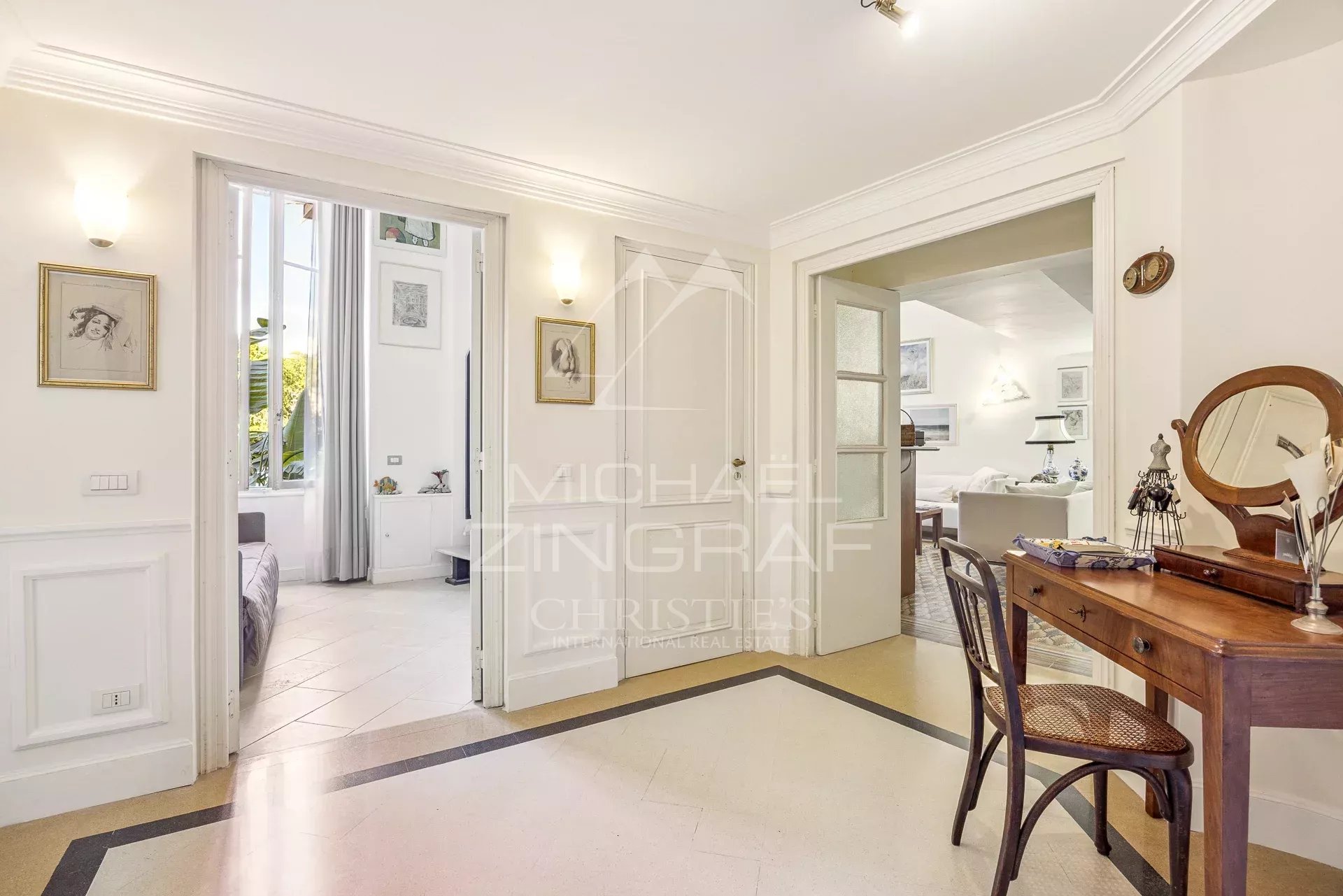 Bright hallway with a wooden dressing table and round mirror on the right, two open doorways leading to bedrooms and living area, framed art on the walls.
