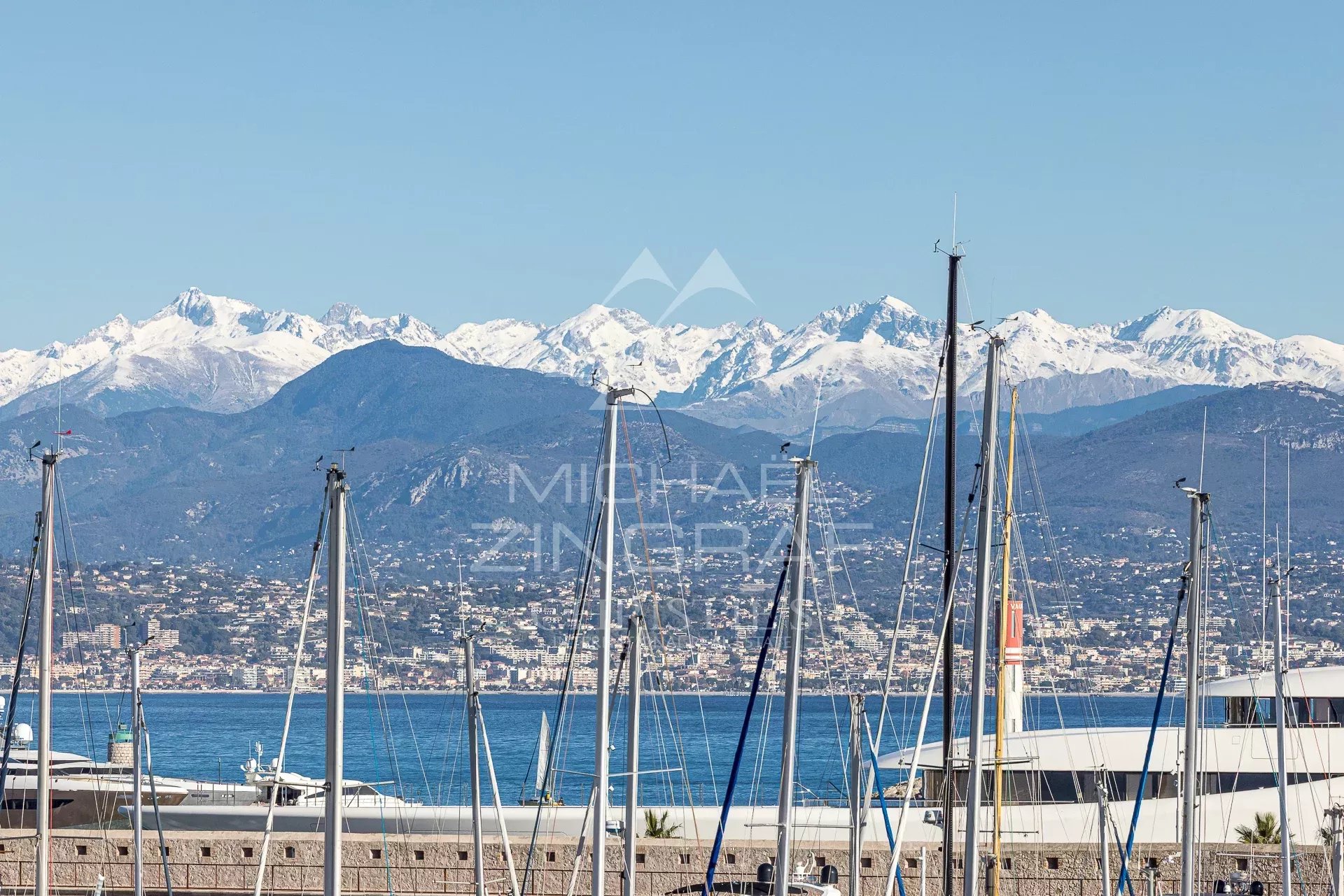 Vue panoramique des montagnes et du port d'Antibes avec des voiliers au premier plan.
