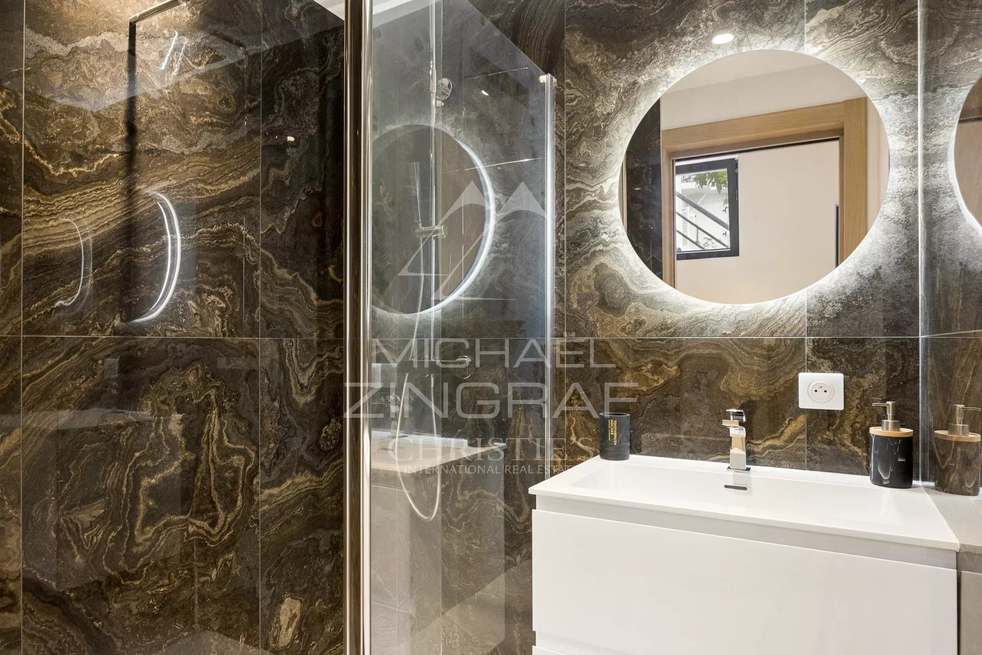 Salle de bain moderne dans un duplex à Antibes Salle de bain moderne avec douche, miroir rond et meuble lavabo en blanc.