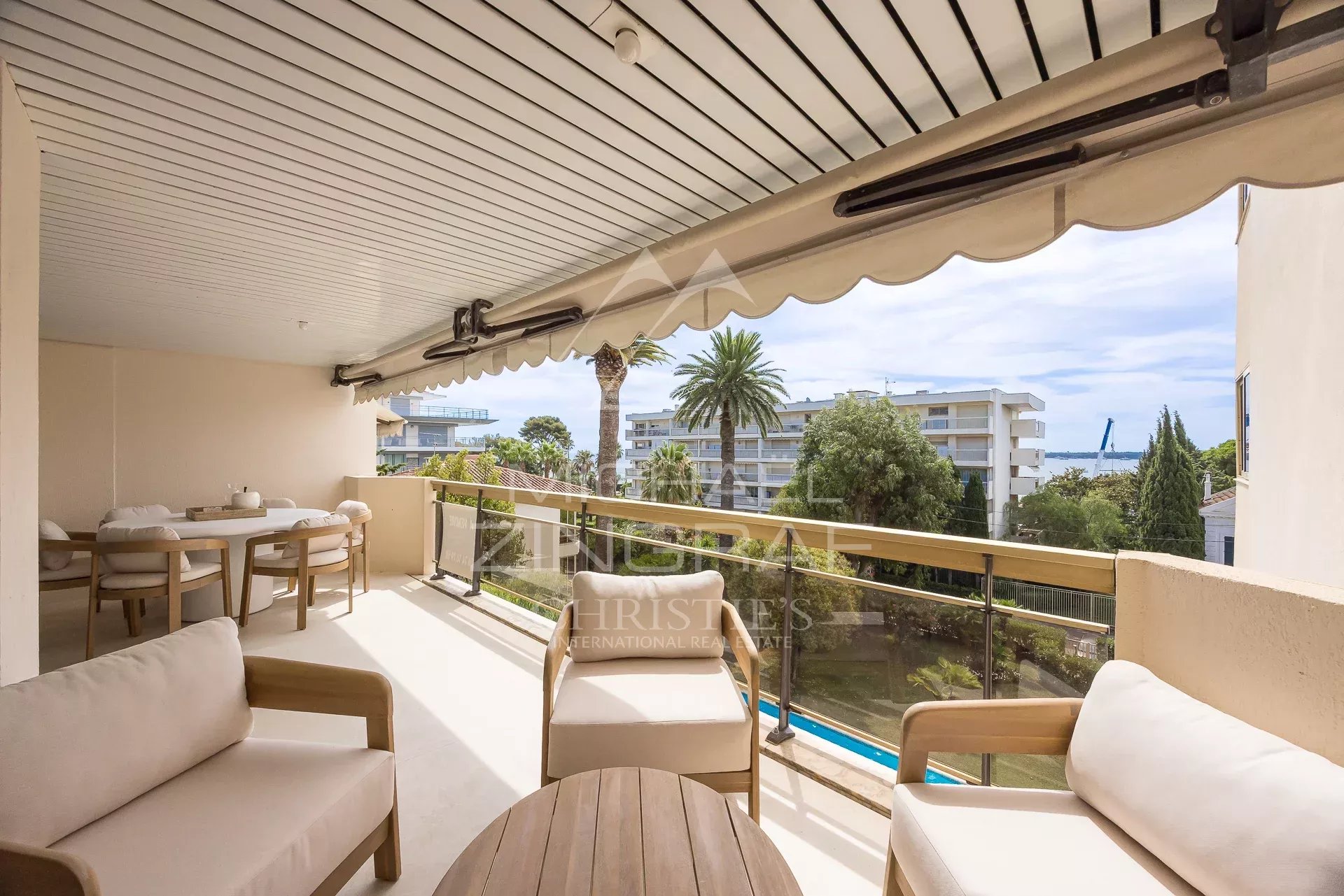 Balcony seating area with beige cushioned chairs and a round table, overlooking palm trees and a coastal neighborhood.