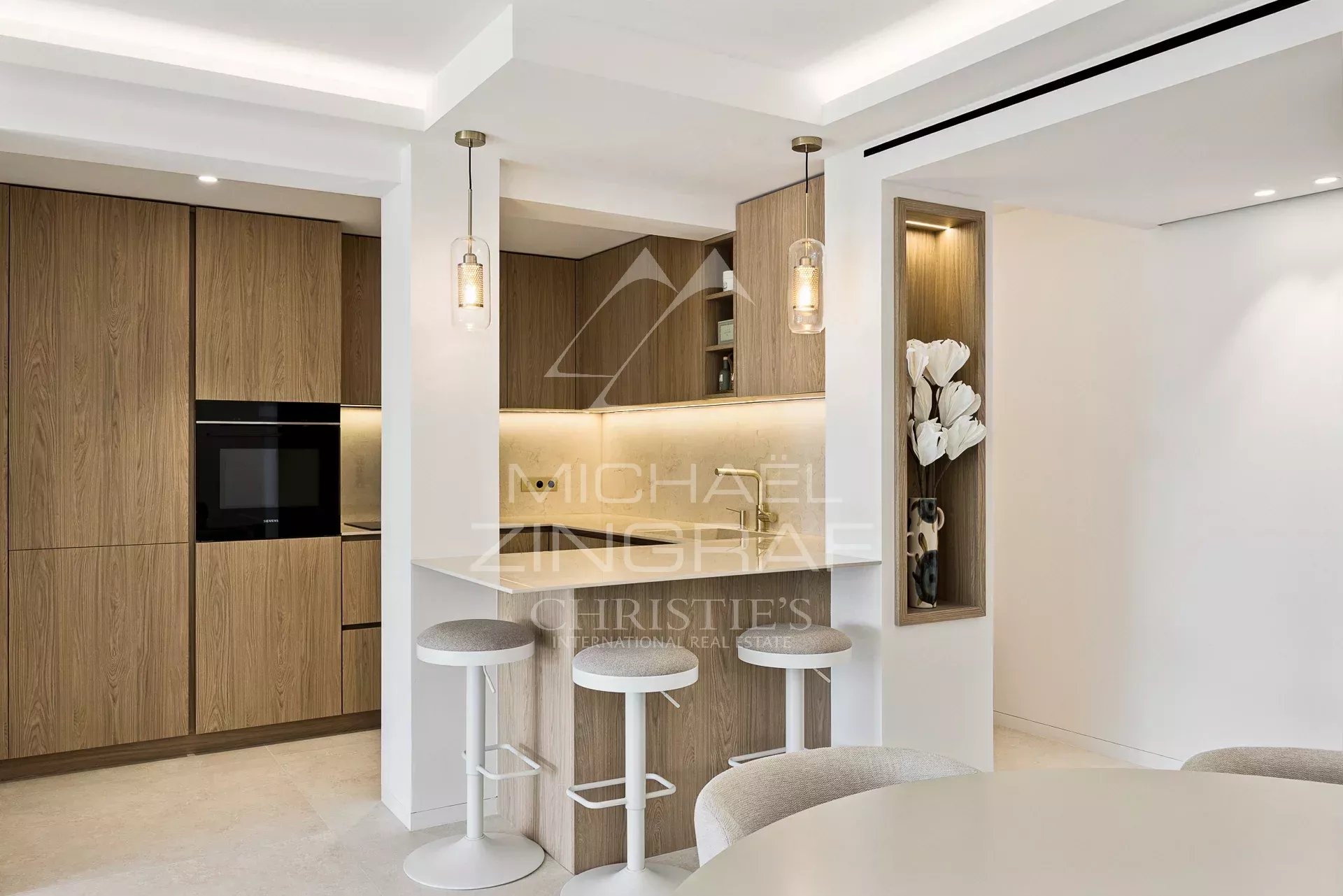 Modern kitchen with wood veneer cabinets, a built-in oven, and a white island with three bar stools under pendant lights.