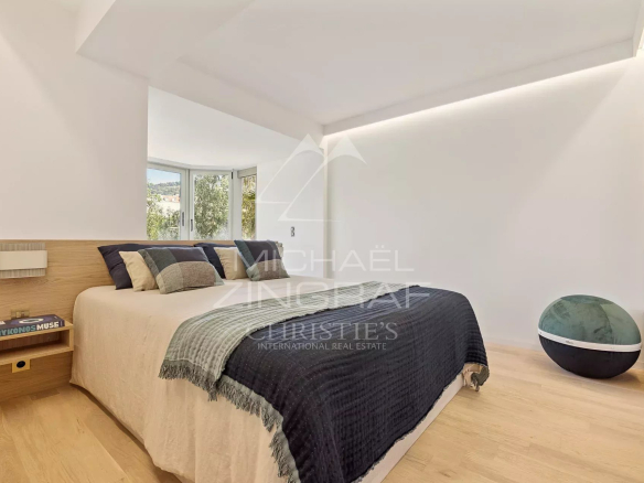 Minimalist bedroom with a low wooden headboard, cream bedding, and a dark quilt; a curved window view and a green pouf on the right side of the room.