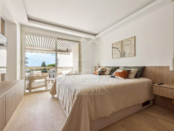 Bright hotel-style bedroom with a large bed, beige bedding, and a balcony door opening to an outdoor seating area and ocean view.