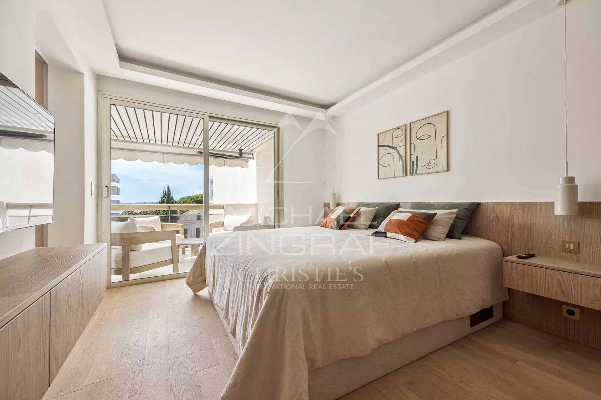 Bright hotel-style bedroom with a large bed, beige bedding, and a balcony door opening to an outdoor seating area and ocean view.