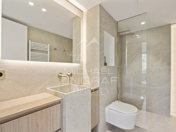 Modern beige bathroom with a wall-mounted sink, wooden vanity, backlit mirror, and a glass-enclosed shower beside a wall-hung toilet.