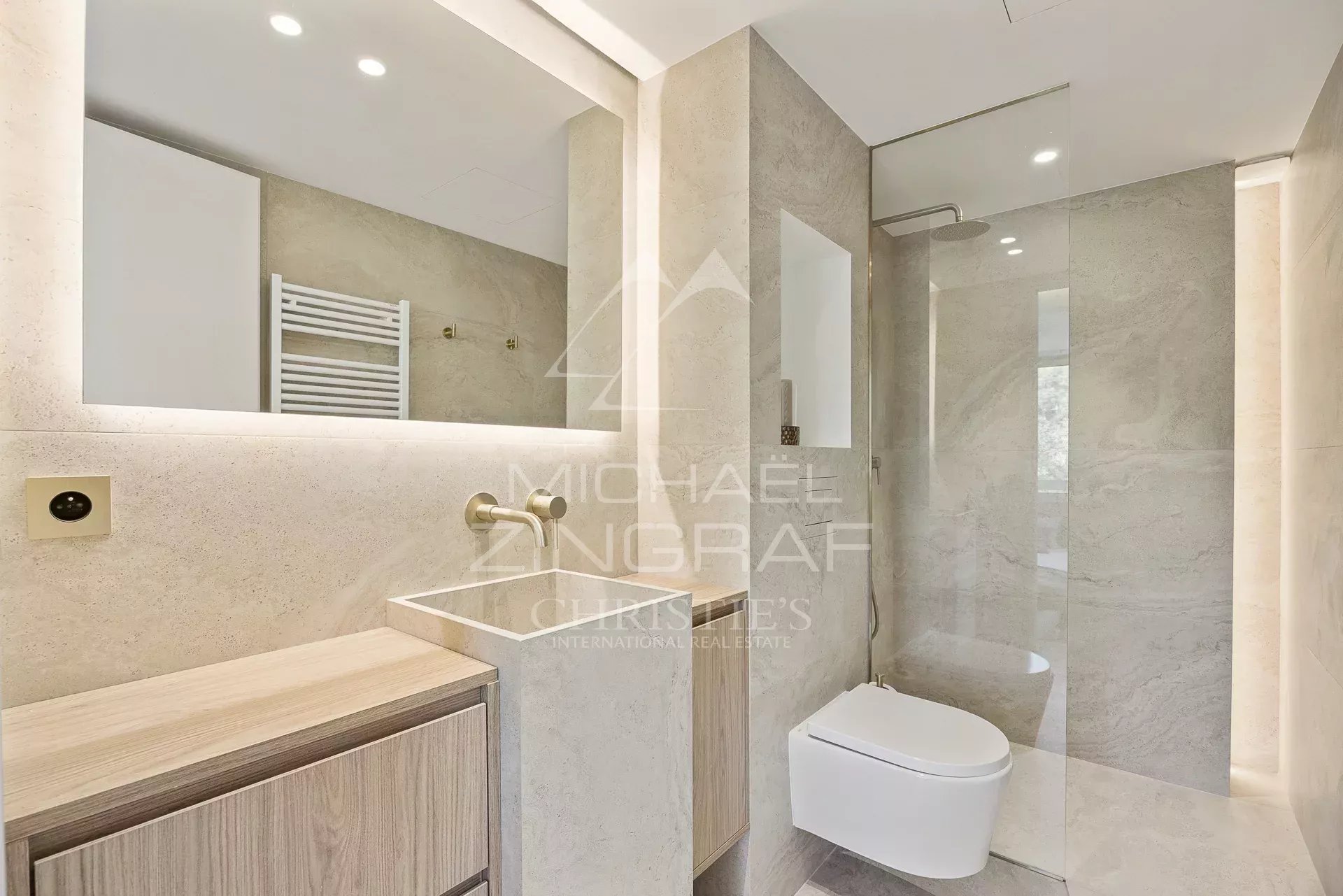 Modern beige bathroom with a wall-mounted sink, wooden vanity, backlit mirror, and a glass-enclosed shower beside a wall-hung toilet.