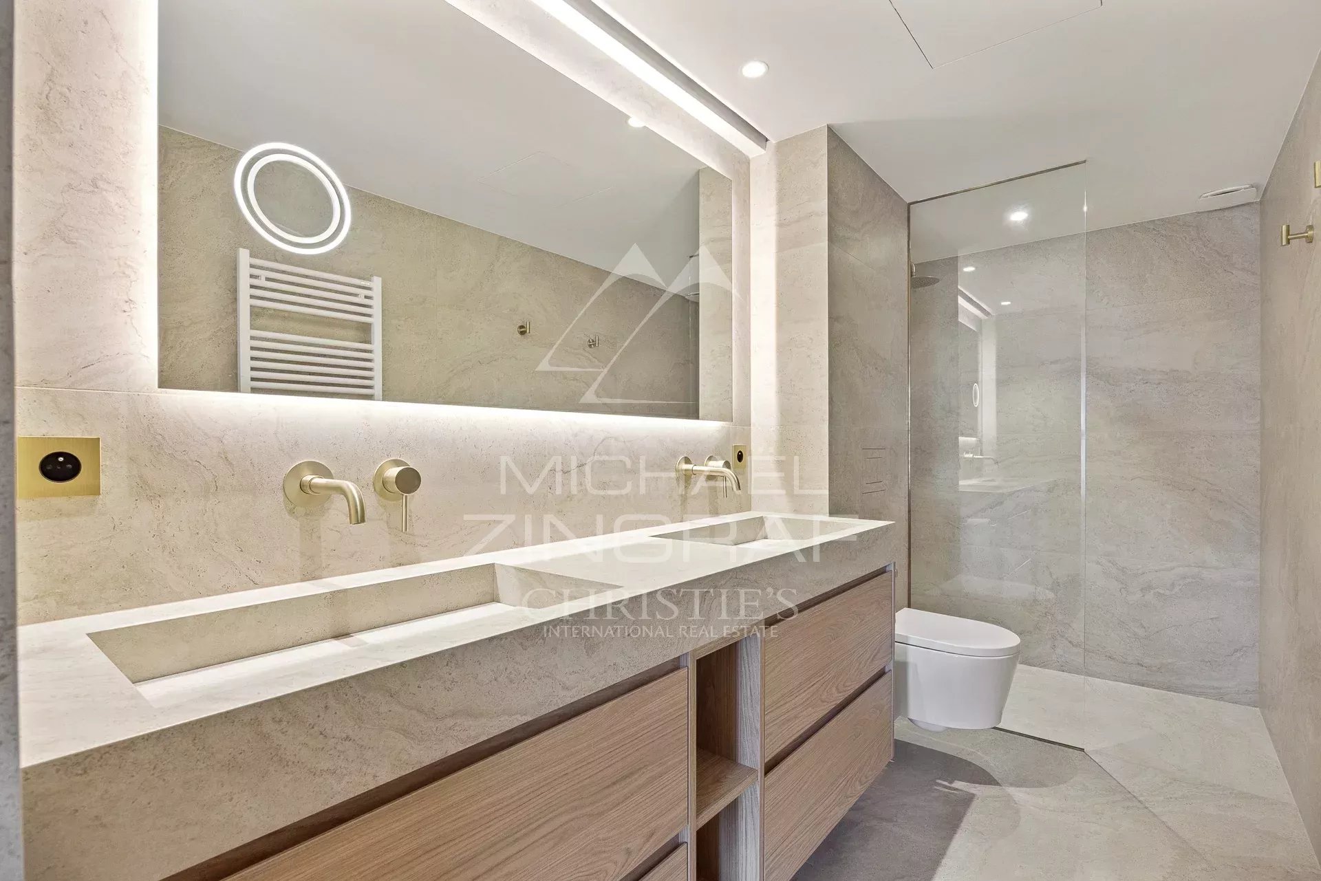 Contemporary bathroom with a double-sink vanity, light wood drawers, and a glass-walled shower in neutral marble tones.