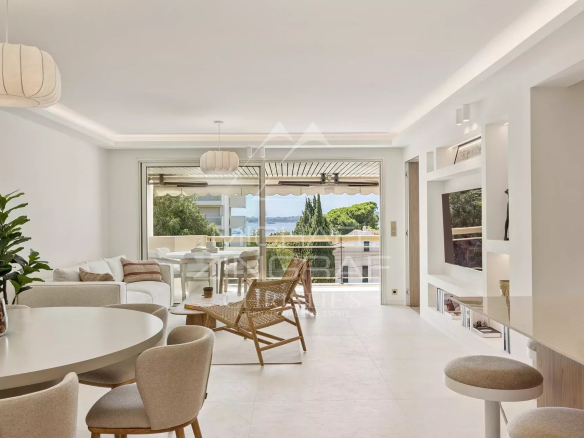 Bright open-plan living room with white furniture, round dining table, wicker lounge chair, and glass doors to a balcony with a sea view.