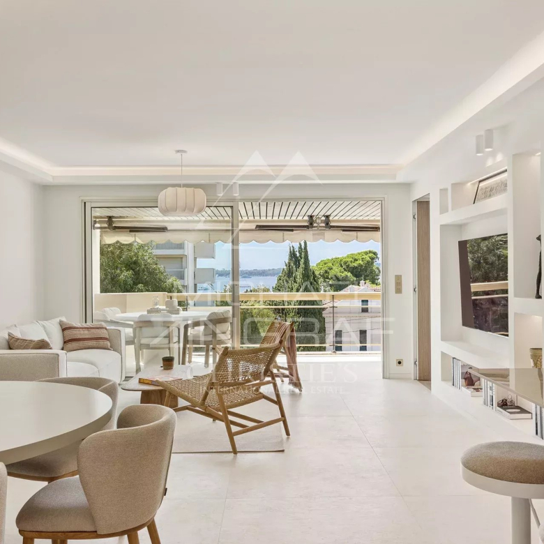 Bright open-plan living room with white furniture, round dining table, wicker lounge chair, and glass doors to a balcony with a sea view.