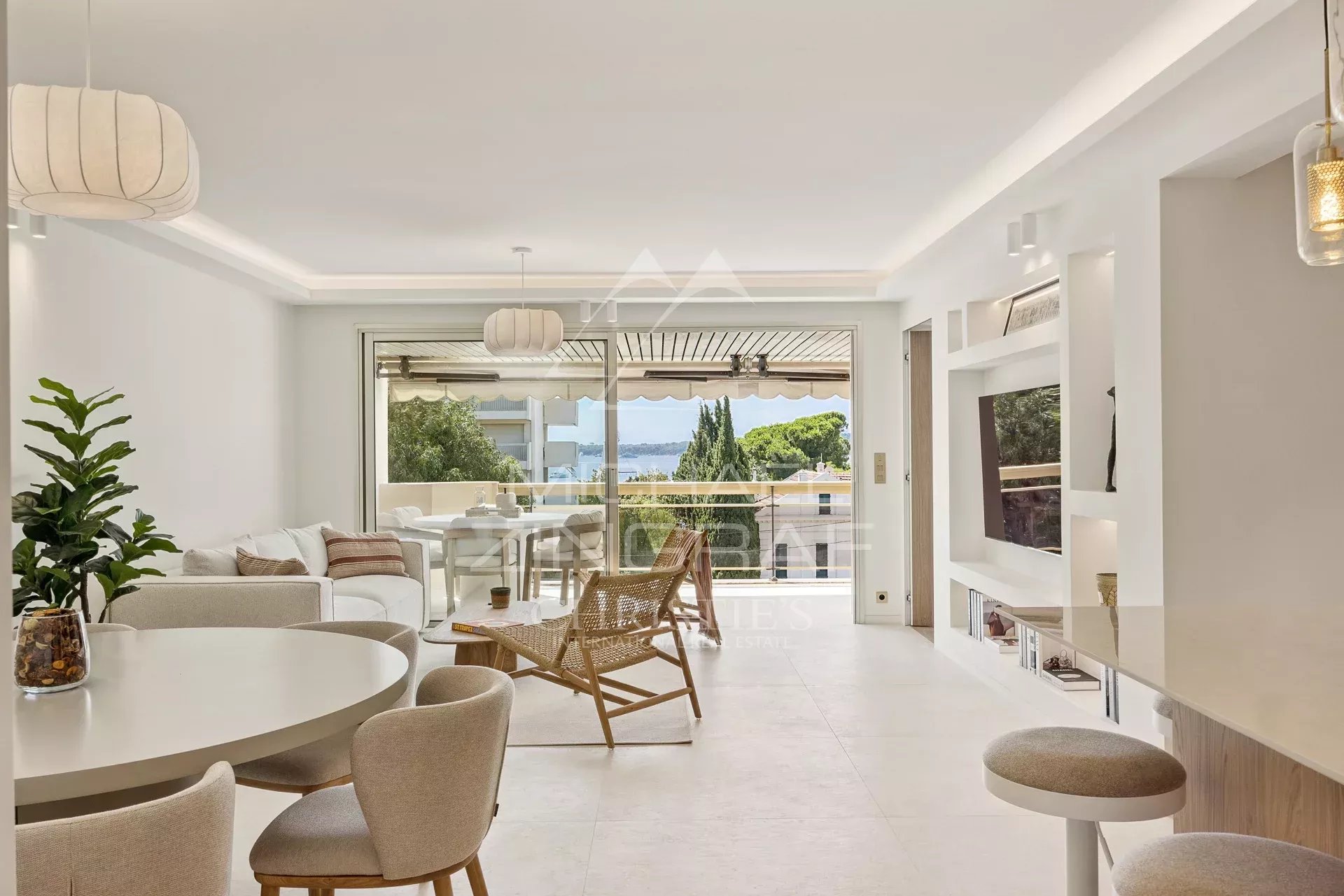 Bright open-plan living room with white furniture, round dining table, wicker lounge chair, and glass doors to a balcony with a sea view.
