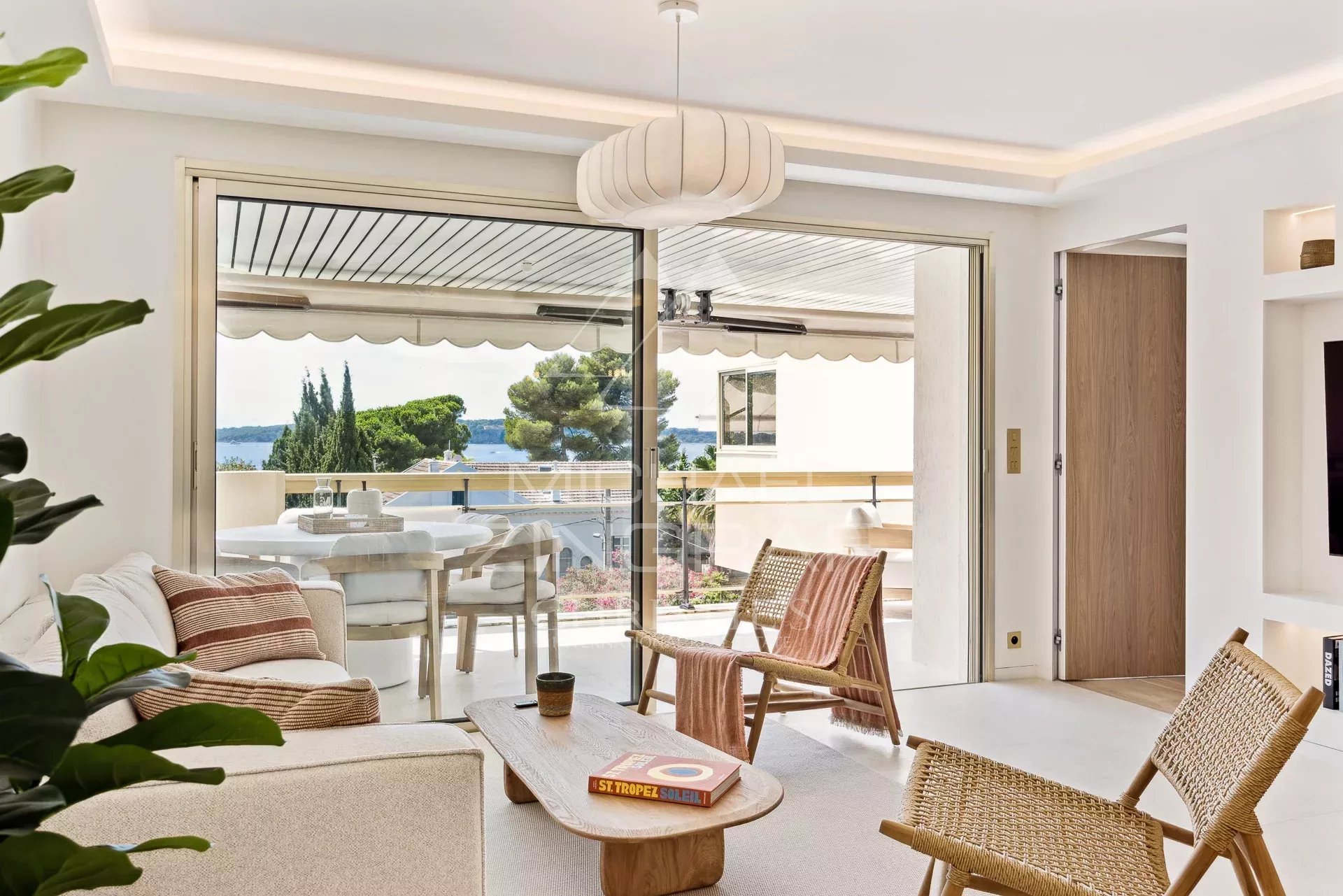 Bright living room with sliding glass doors opening to a balcony overlooking trees and the sea; wicker chairs and a wooden coffee table indoors.