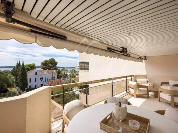 Sunlit balcony with round white table and cushioned chairs, overlooking a coastal neighborhood and the sea.