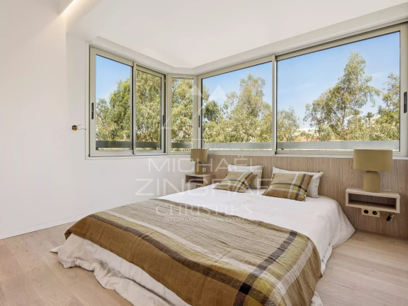Bright bedroom with a large bay of windows overlooking trees, a low wood headboard, and a bed covered in a plaid brown-and-beige blanket.