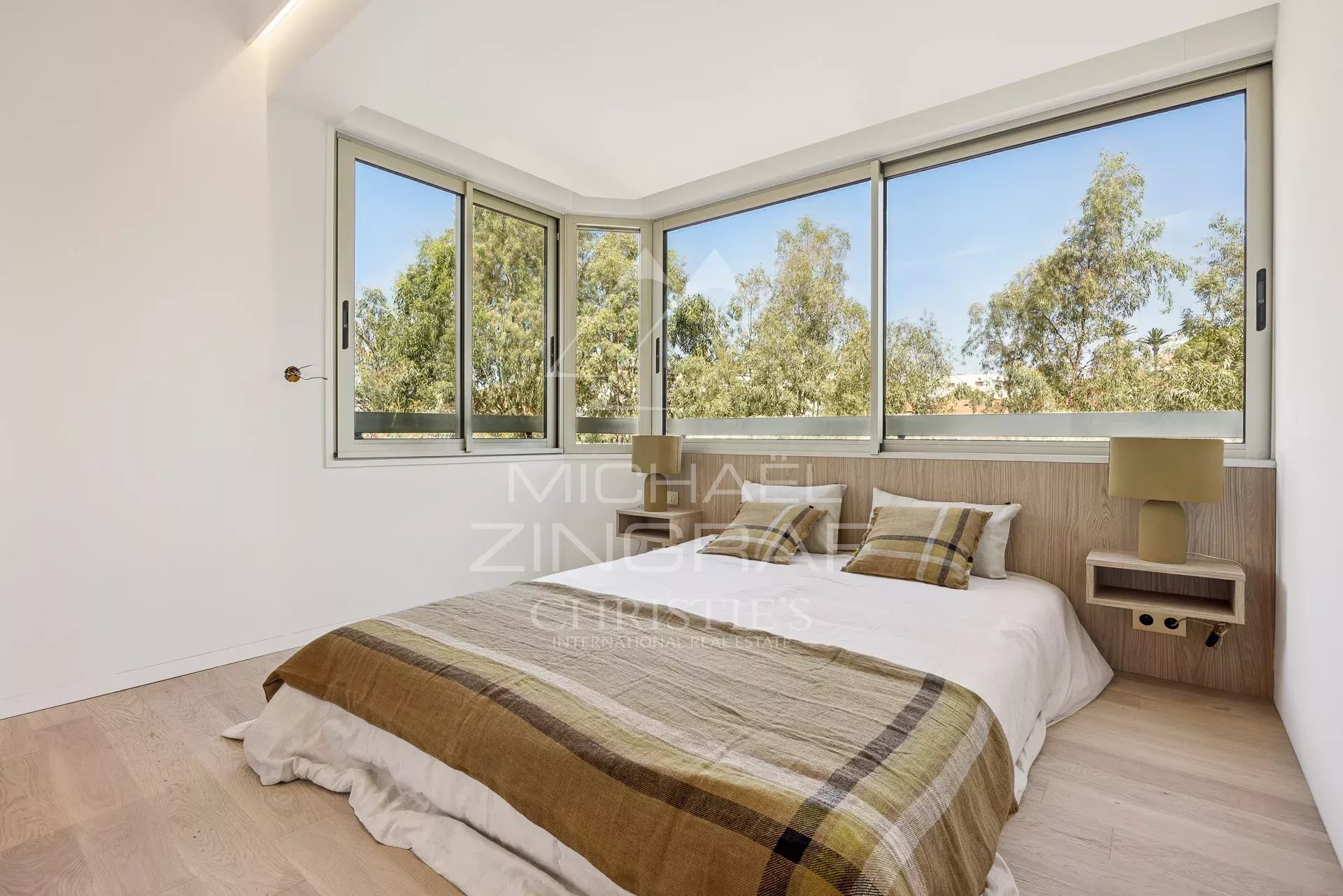 Bright bedroom with a large bay of windows overlooking trees, a low wood headboard, and a bed covered in a plaid brown-and-beige blanket.