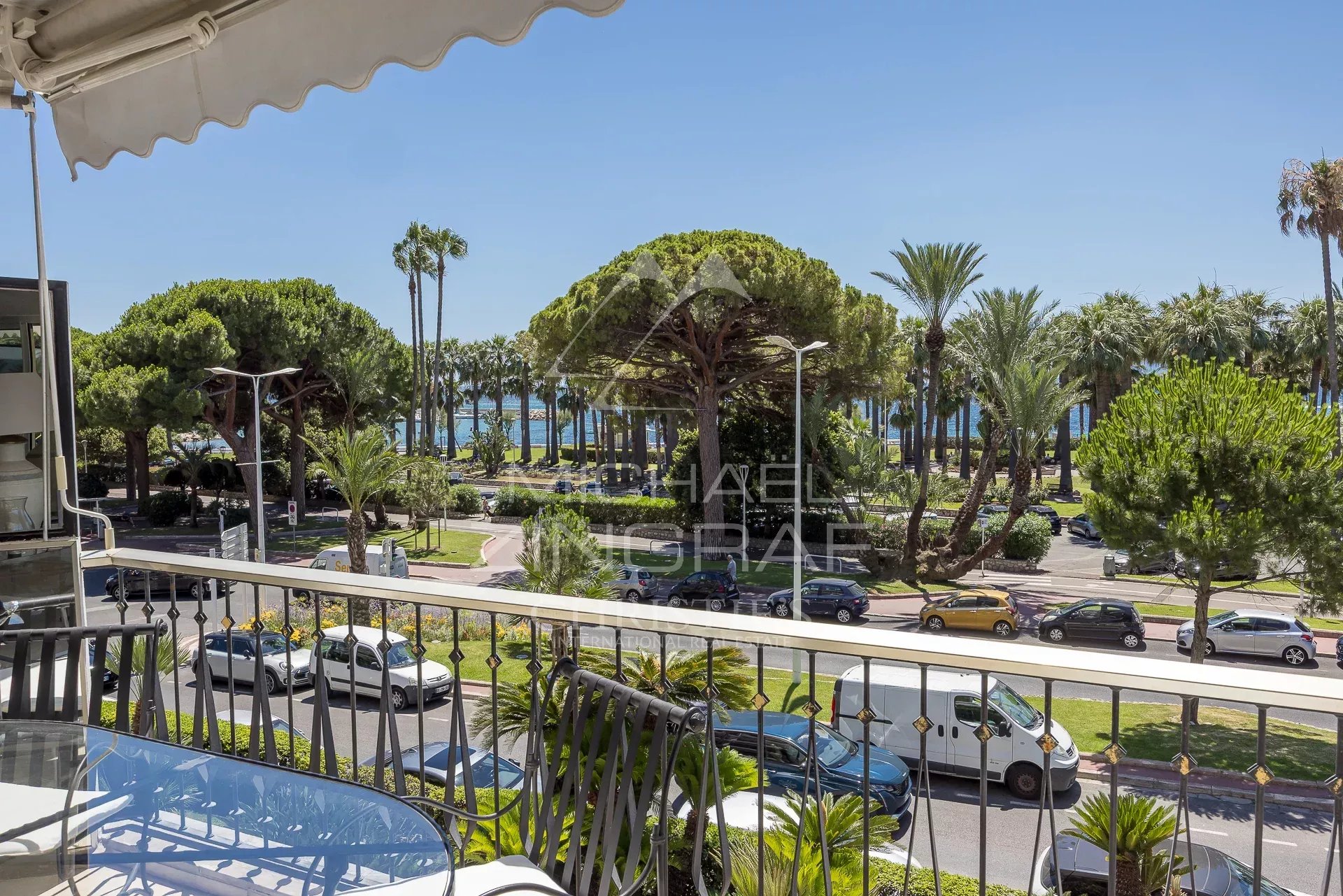 Vue depuis une terrasse d'un appartement à Cannes, avec des palmiers et la Croisette en arrière-plan.