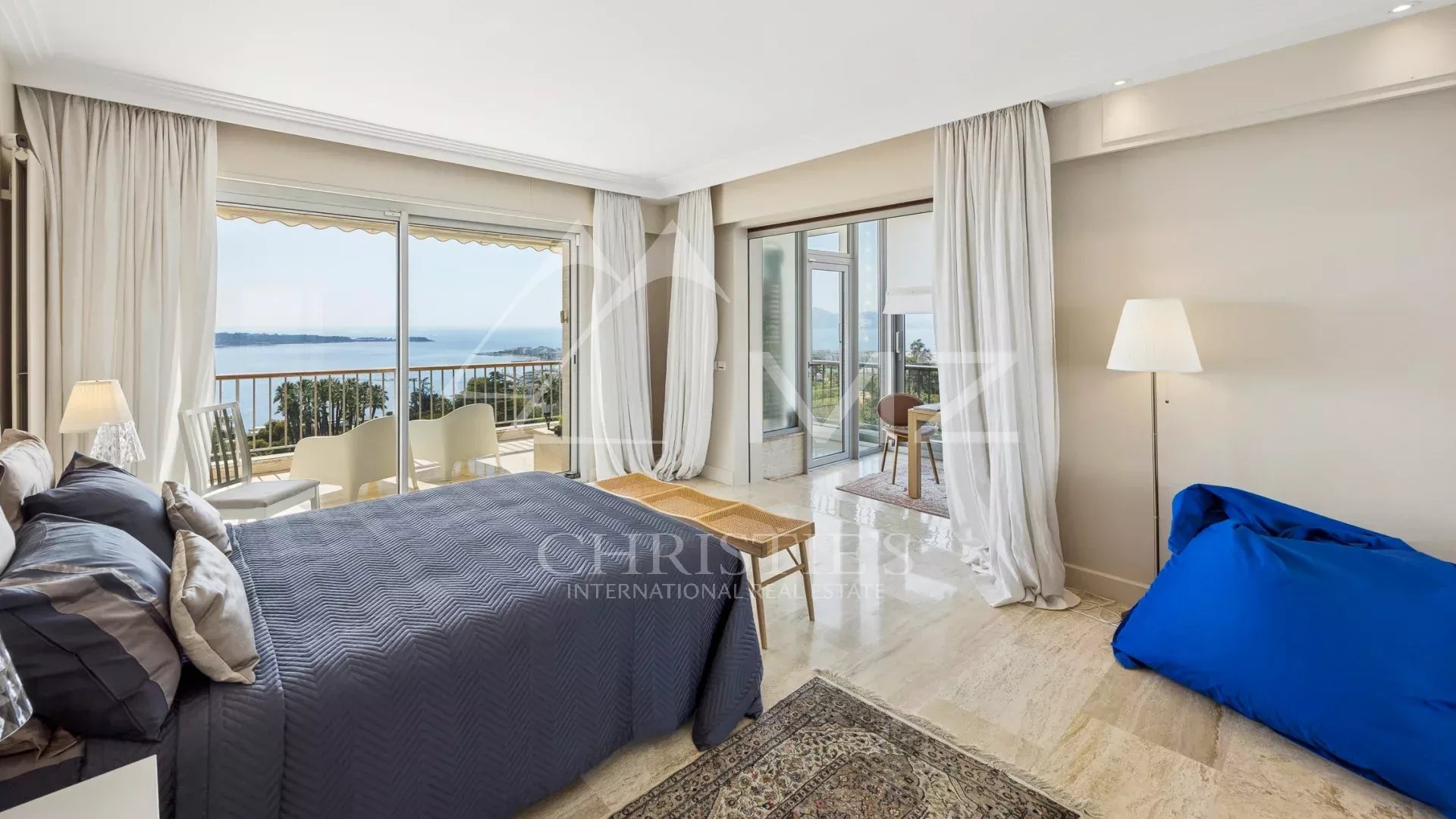 Bright hotel bedroom with a navy quilted bed, white drapes, and a balcony overlooking the sea.