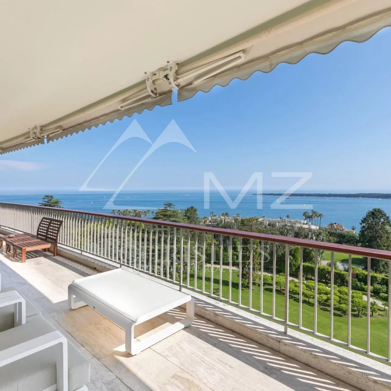 Balcony with white lounge chairs and a coffee table overlooking the blue sea and distant shoreline on a sunny day.
