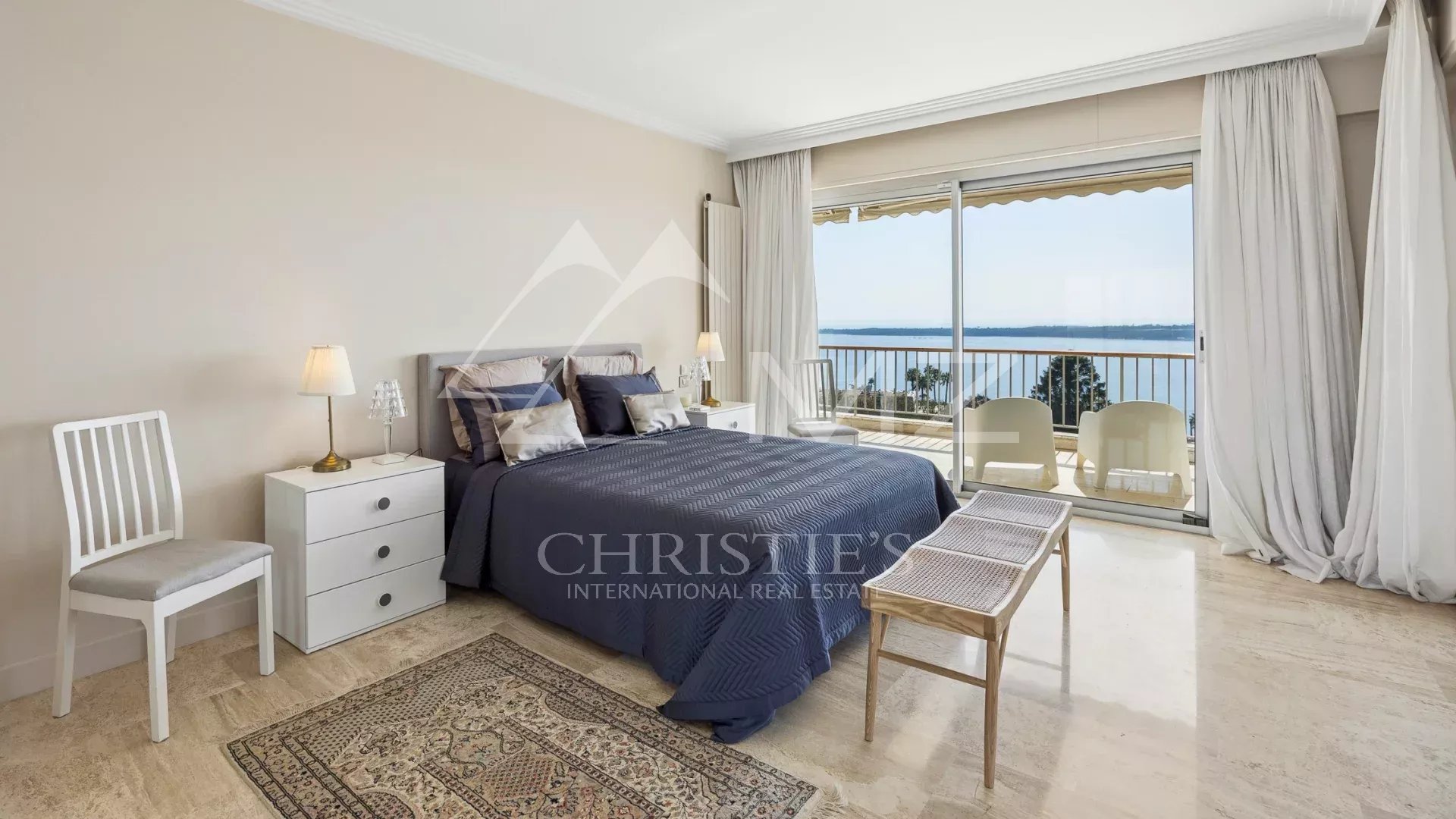 Bedroom with a queen bed, navy cover, and white nightstands facing a sea-view balcony.