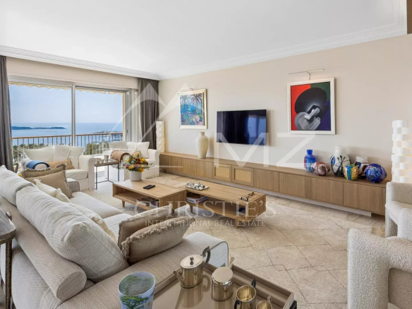 Bright living room with a sea view through glass doors, white sectional sofa, wooden coffee table, and a long wall cabinet with art and vases.