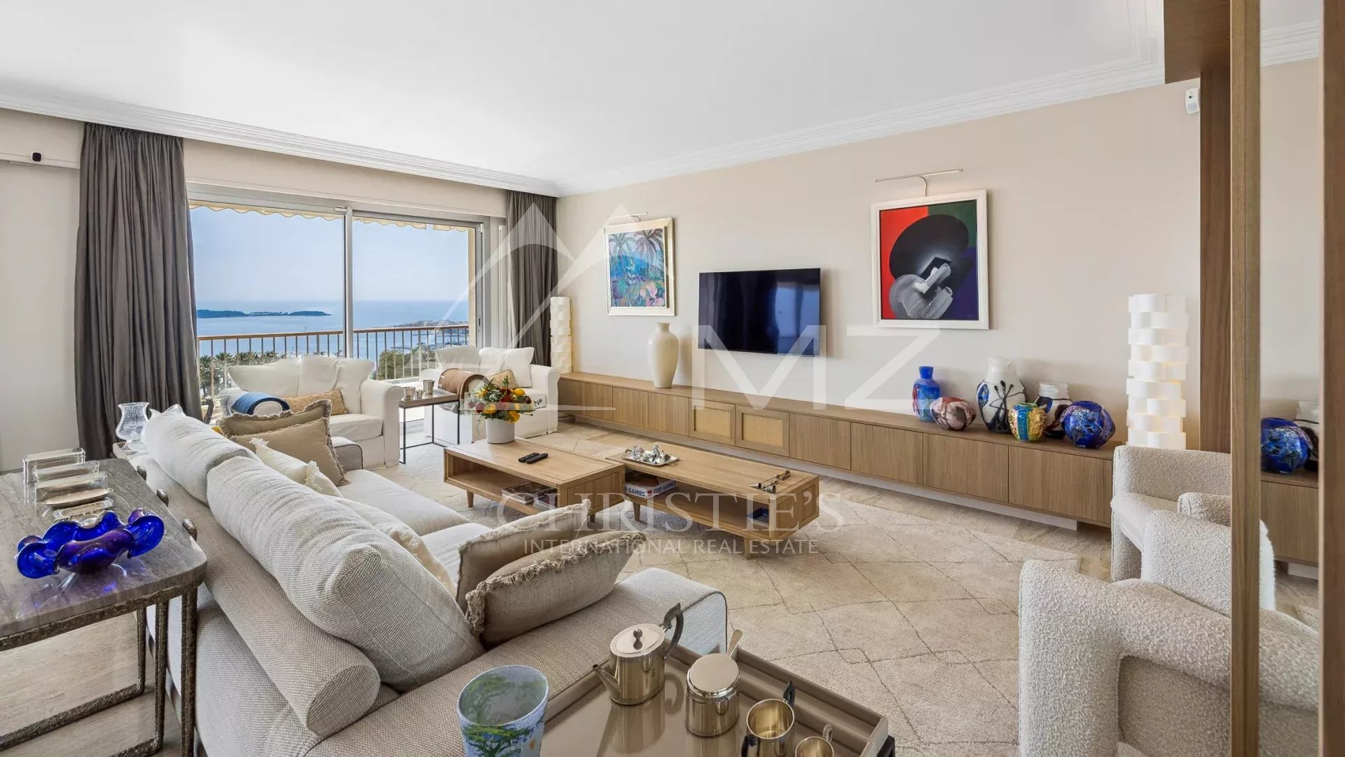 Bright living room with a sea view through glass doors, white sectional sofa, wooden coffee table, and a long wall cabinet with art and vases.