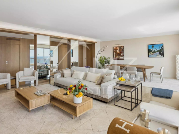 Bright open-plan living room with beige sofas, wooden coffee tables, a dining area, and a balcony view behind a glass wall.
