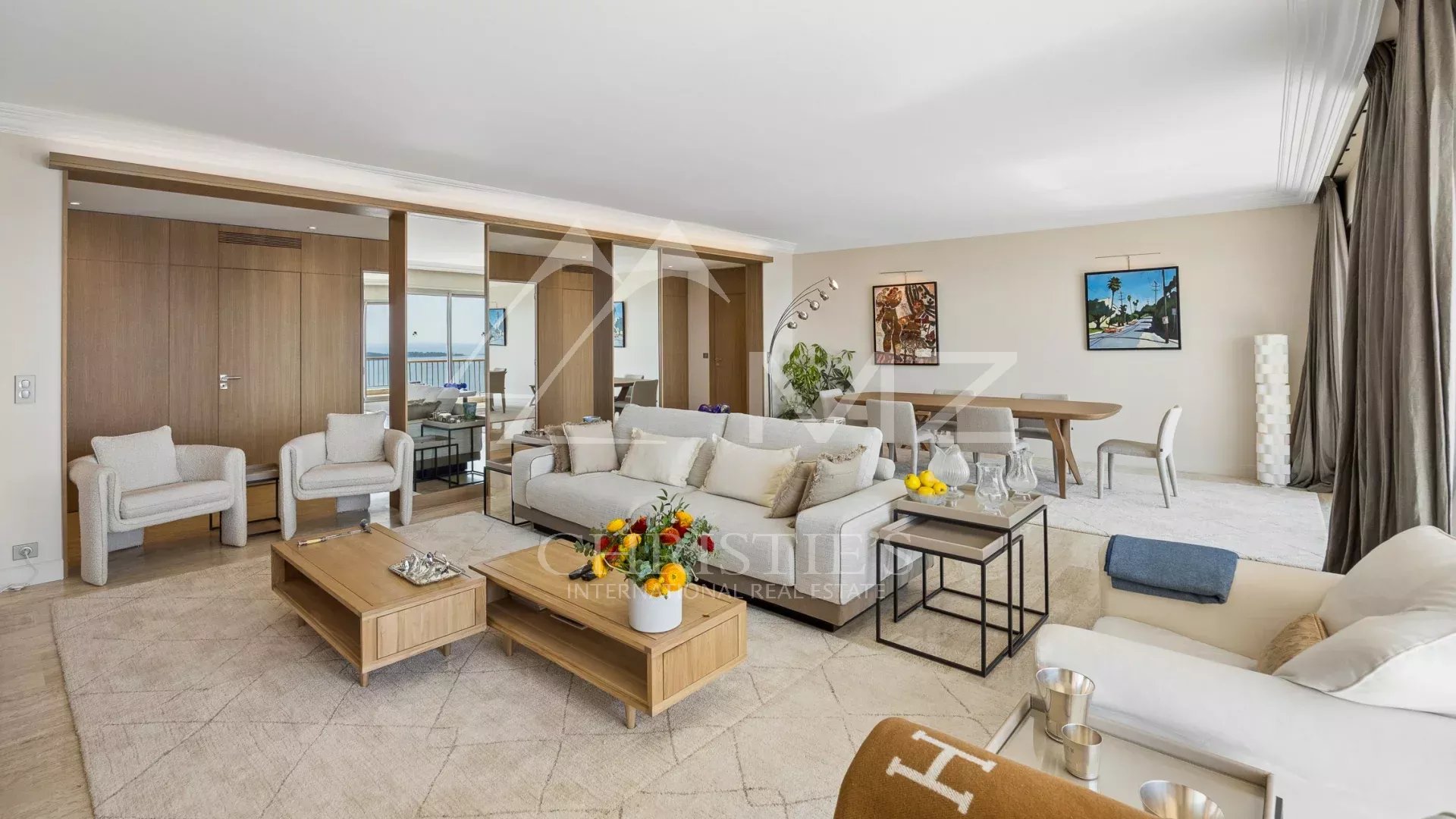 Bright open-plan living room with beige sofas, wooden coffee tables, a dining area, and a balcony view behind a glass wall.