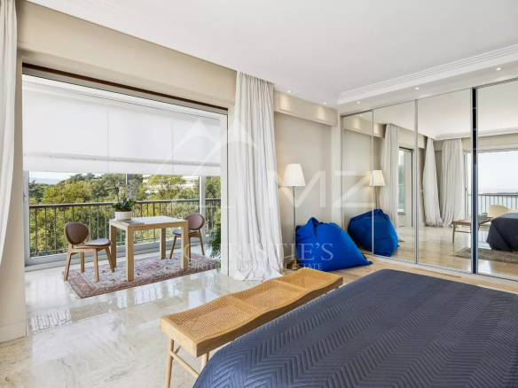 Bright hotel bedroom with balcony seating area, view of trees and water, cream curtains, and mirrored wardrobe door reflection.
