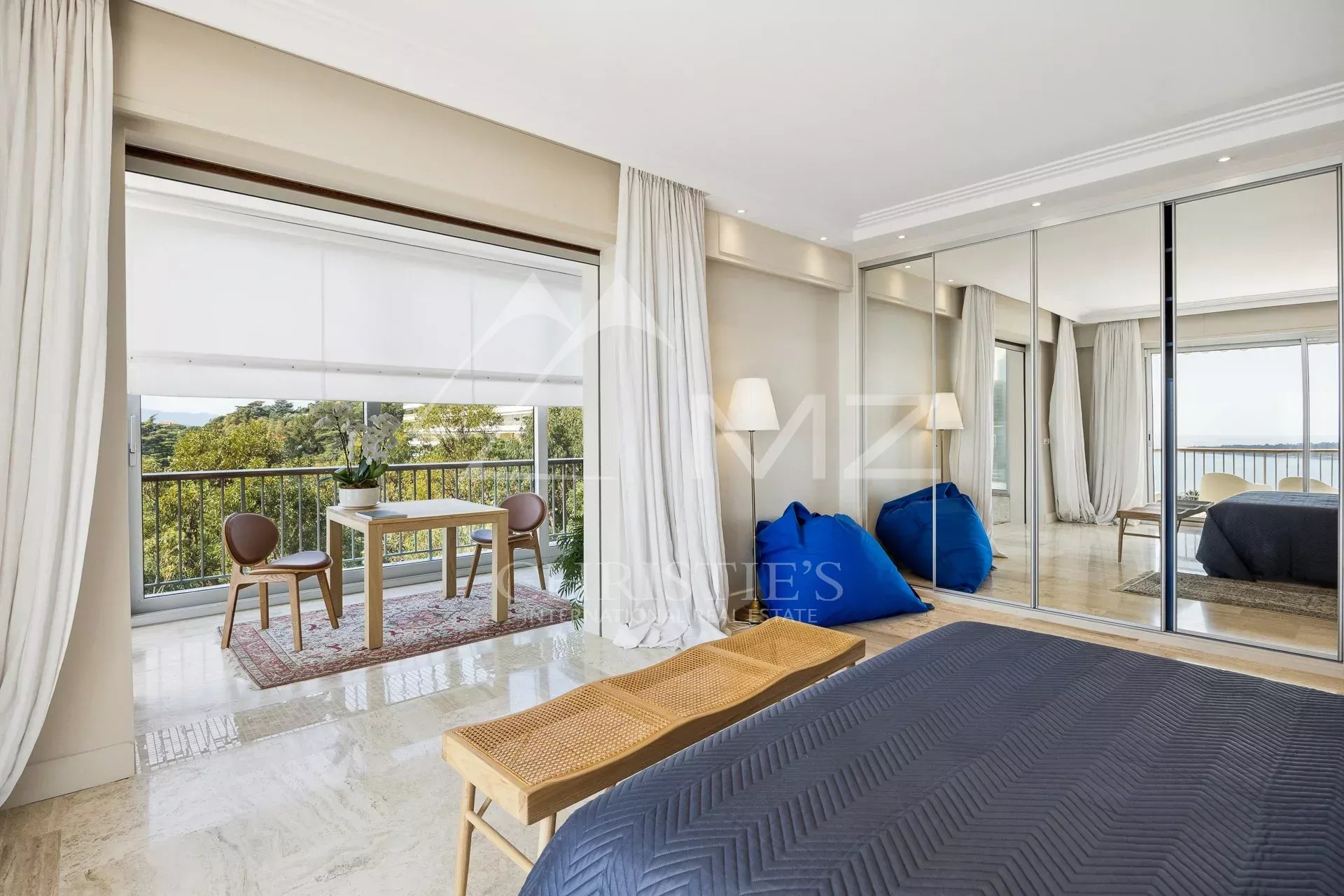 Bright hotel bedroom with balcony seating area, view of trees and water, cream curtains, and mirrored wardrobe door reflection.