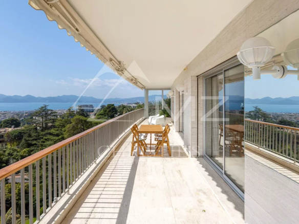 Balcony with wooden table and chairs overlooking a coastal city and distant mountains under a clear blue sky.