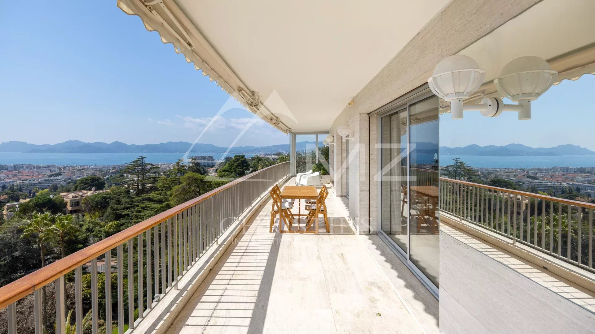 Balcony with wooden table and chairs overlooking a coastal city and distant mountains under a clear blue sky.