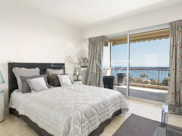 Bedroom with a gray upholstered bed, white quilt, and multiple pillows, beside glass doors to a balcony overlooking the sea.