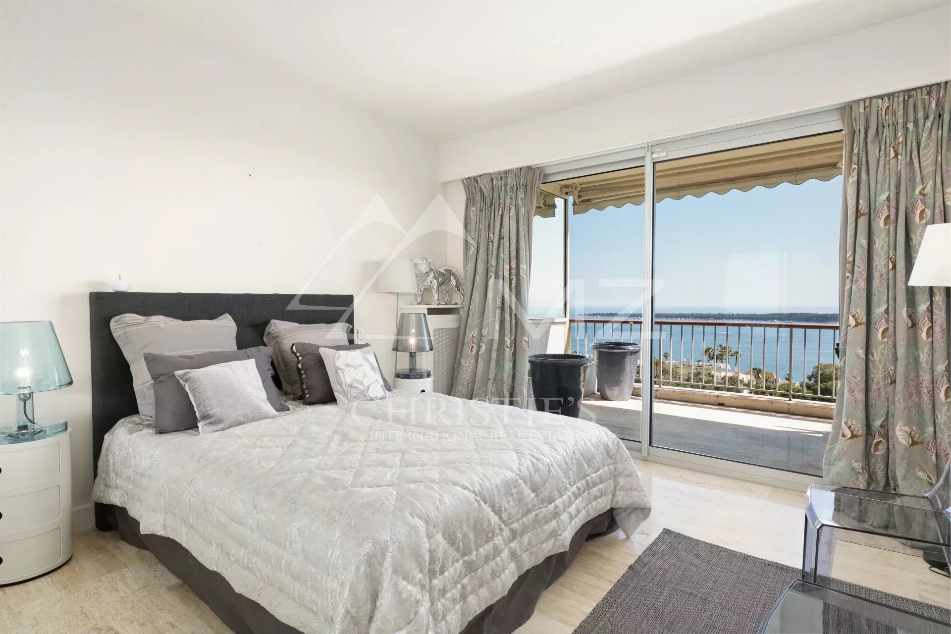 Bedroom with a gray upholstered bed, white quilt, and multiple pillows, beside glass doors to a balcony overlooking the sea.