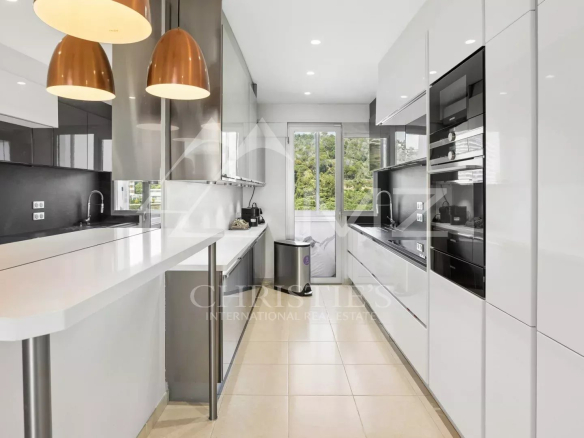Modern white kitchen with a long island, copper pendant lights, black backsplash, and built-in ovens along the right wall.