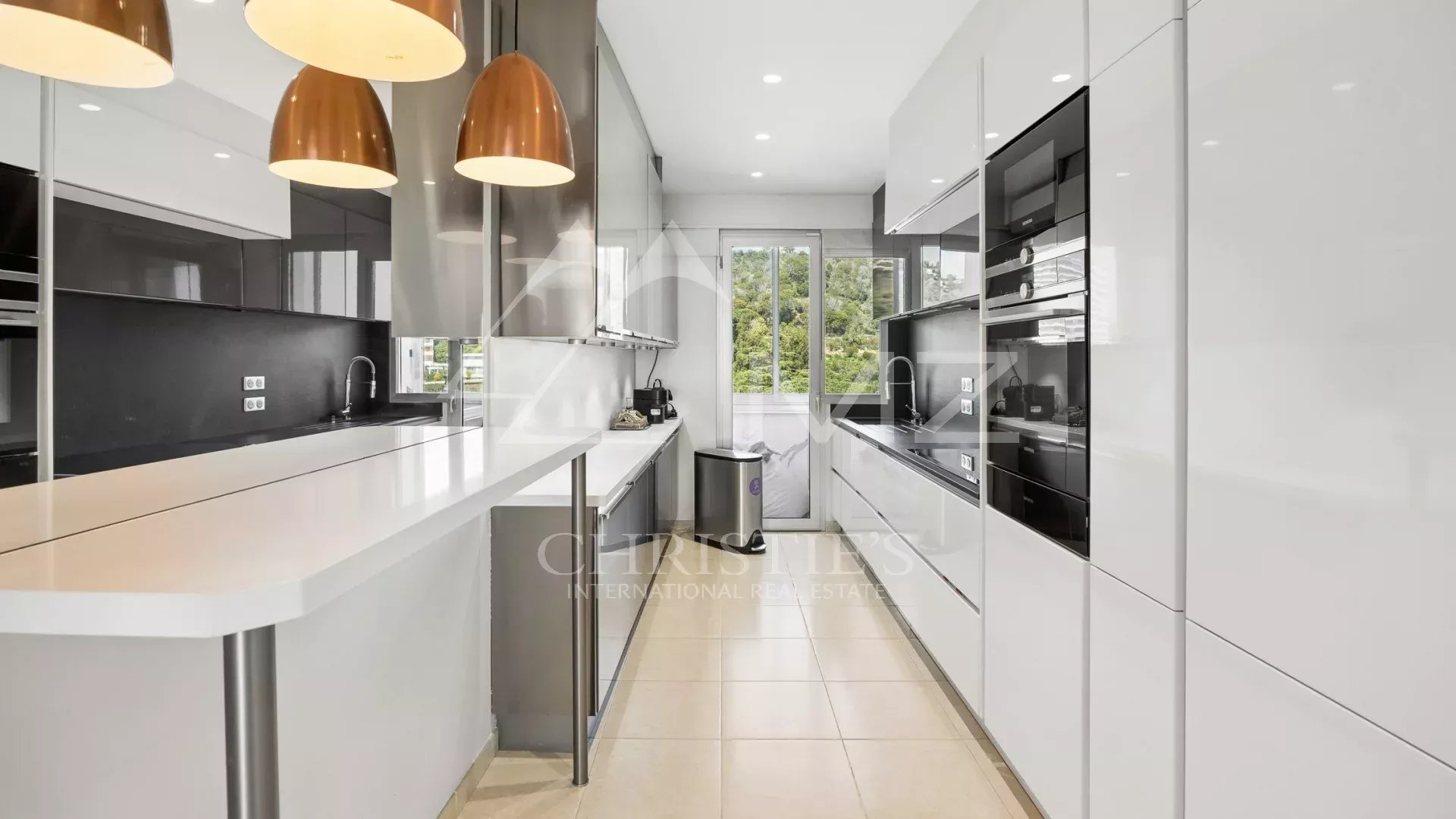 Modern white kitchen with a long island, copper pendant lights, black backsplash, and built-in ovens along the right wall.