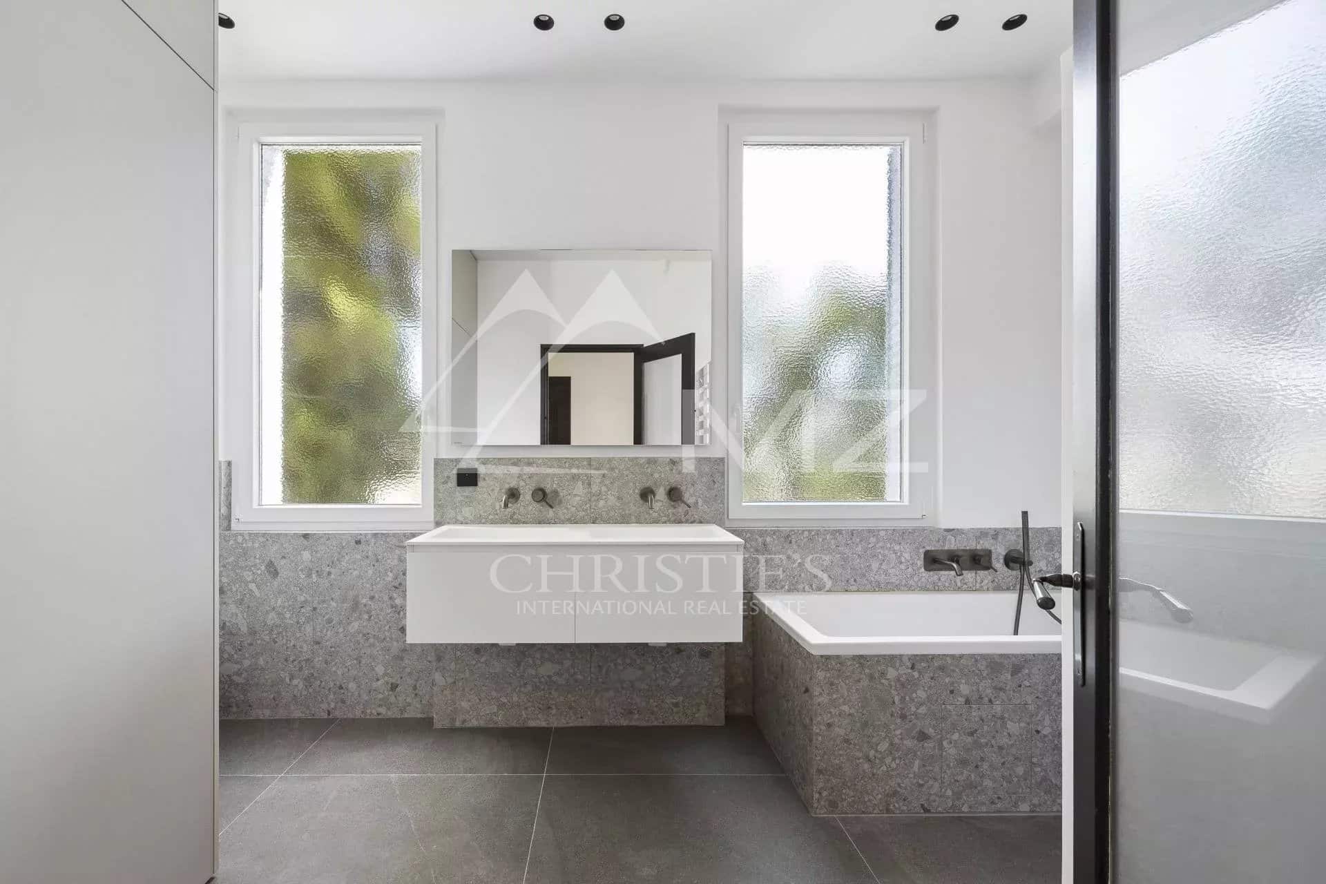 Salle de bains moderne avec baignoire et vasque en pierre, fenêtres avec vue sur la verdure.
