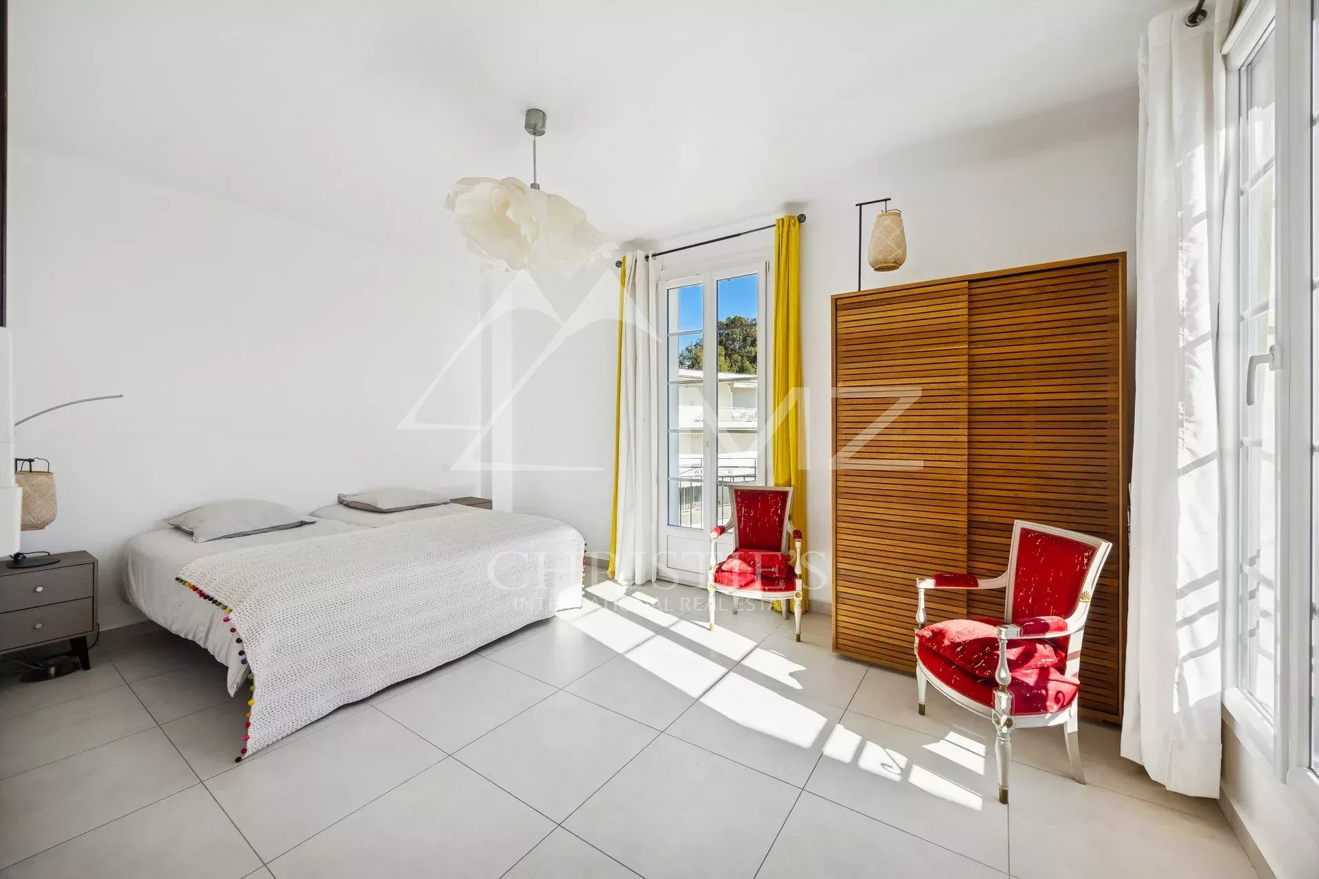 Chambre d’appartement à Cannes Chambre lumineuse avec lit double et fauteuils rouges, fenêtres ouvertes sur la vue.