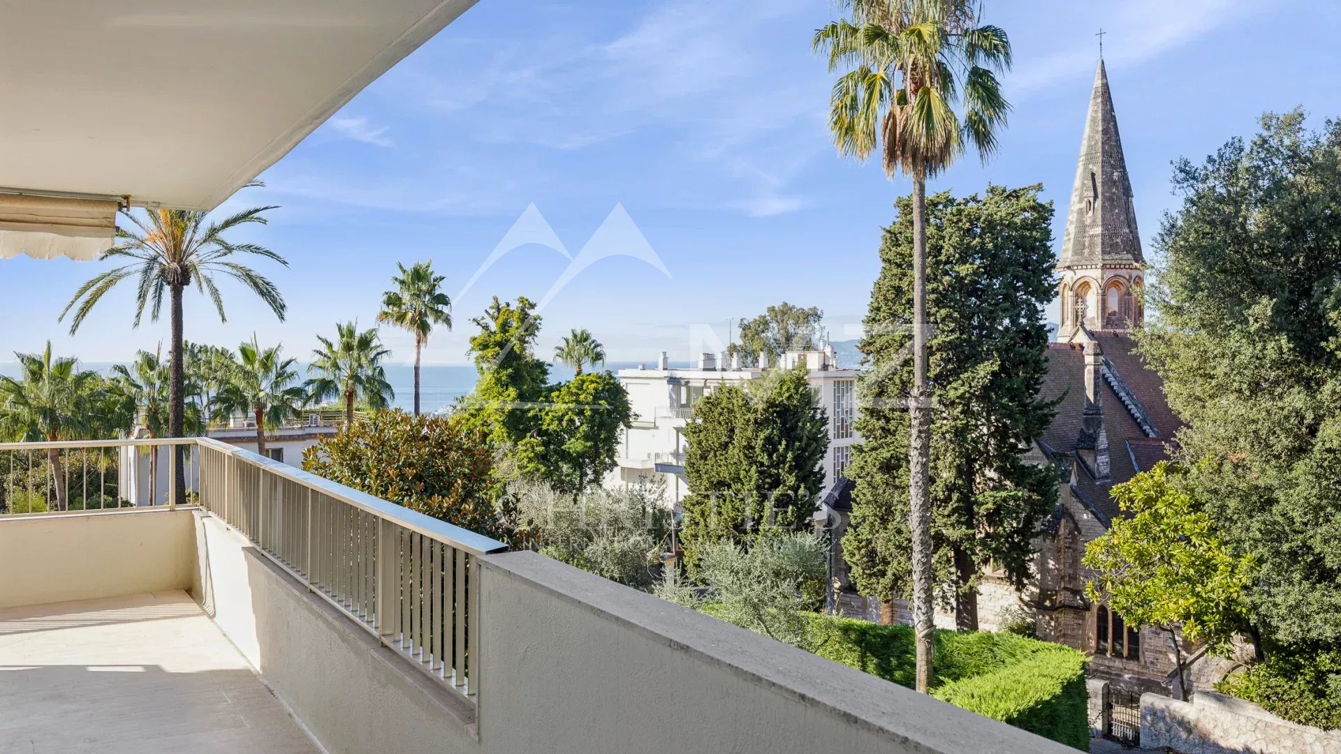 Vue depuis la terrasse d'un appartement à Cannes, avec des palmiers et l'église Saint-Georges en arrière-plan.