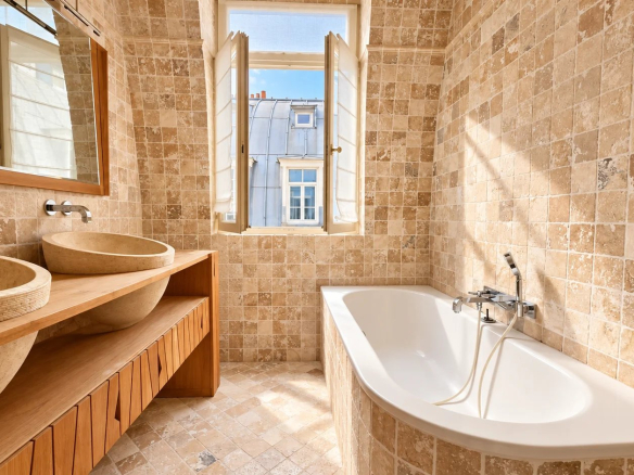 Salle de bain moderne avec baignoire et vasques en pierre, fenêtres ouvertes sur l'extérieur.
